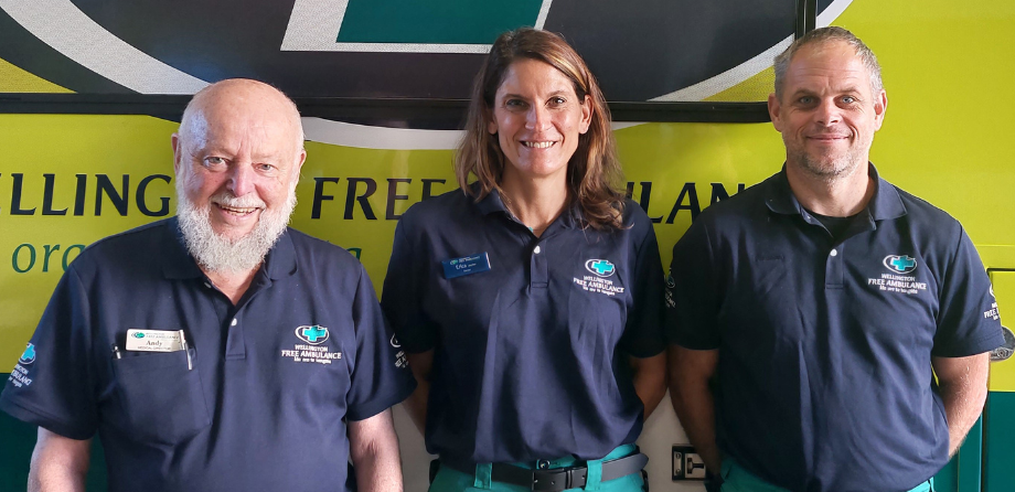 3 people in dark blue polo shirts smile in front of an ambulance