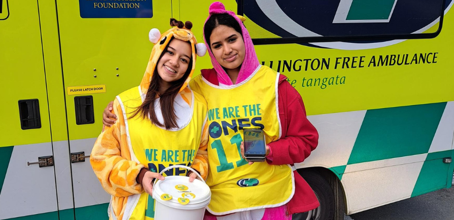 2 people wearing onesies and collector bibs smile in front of an ambulance