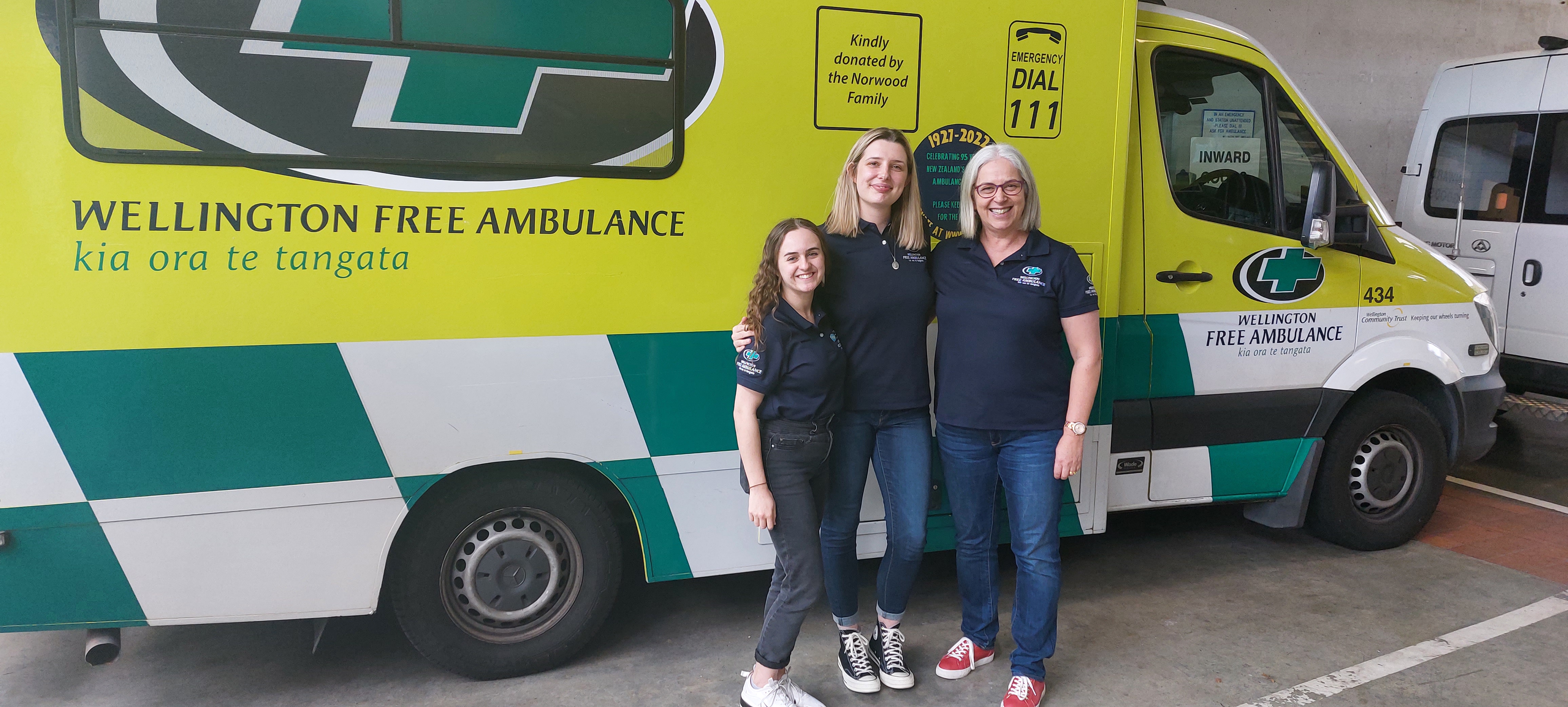 3 women in Wellington Free Ambulance blue polo shirts stand in front of an ambulance