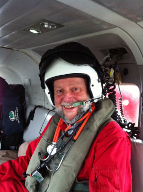A man in a red uniform and helmet smiles from inside a helicopter