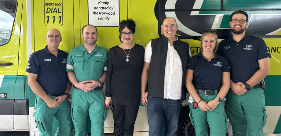 A group of paramedics and a woman in black and a man in a white shirt smile in front of an ambulance