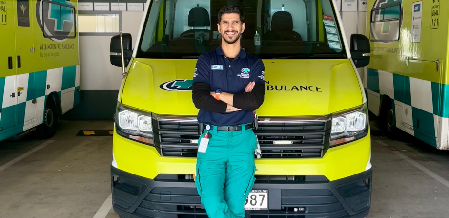 A man in green trousers and a blue top smiles while leaning against an ambulance