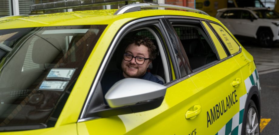 A man in glasses smiles from the front seat of a Wellington Free Ambulance vehicle