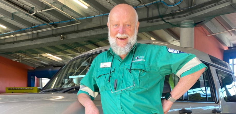 A man in a green uniform leans on a car and smiles