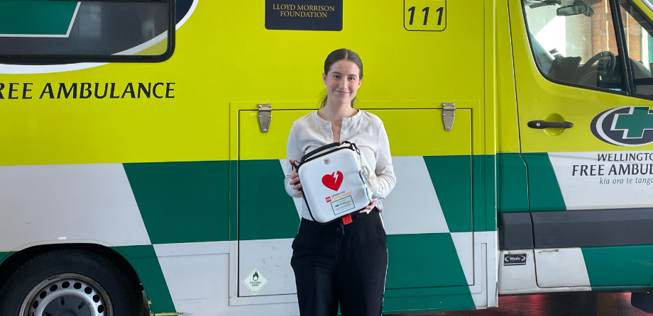 A woman in a white shirt holds an AED and smiles in front of an ambulance