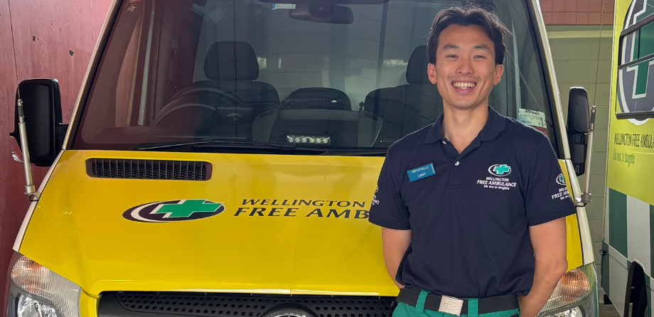 A paramedic in a blue polo shirt smiles in front of an ambulance