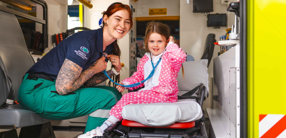A paramedic smiles while a small child in a onesie listens to their chest with a stethoscope