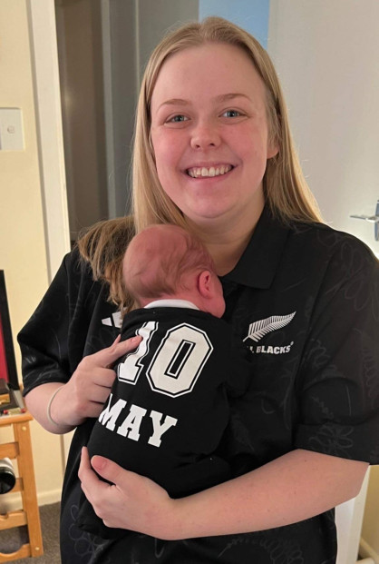 A blonde woman in an All Blacks top holds a baby in an All Blacks top