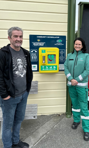 A man and a woman in a green Wellington Free Ambulance uniform stand next to an automated external defibrillator