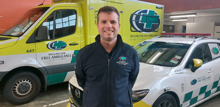 A man in a dark blue jacket smiles in front of Wellington Free Ambulance vehicles