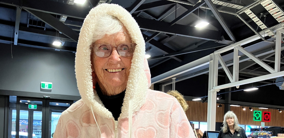 A white haired woman with glasses smiles while wearing a pink onesie