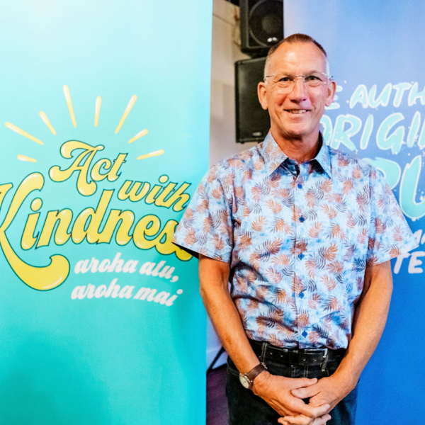A man in a blue shirt and glasses smiles while standing in front of a sign that says Act with Kindness