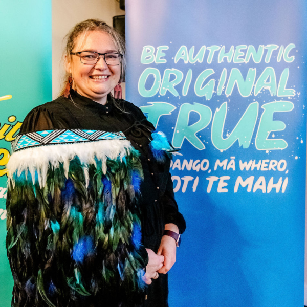 A woman wearing a korowai smiles in front of a sign that says Be authentic, original, true