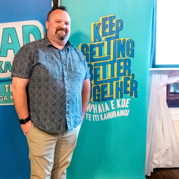 A man in a dark blue shirt smiles in front of a sign that says Keep getting better together
