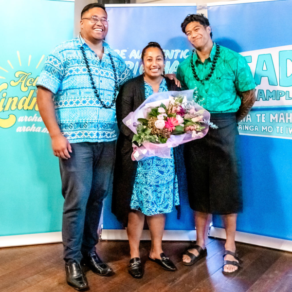 Two men and a woman wearing Pasifika dress smile while holding a bouquet of flowers
