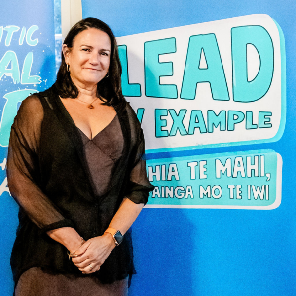 A woman in a brown dress smiles in front of a sign that says Lead by Example