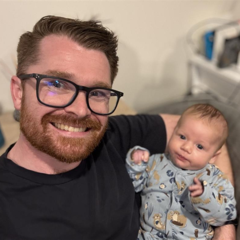 A man in black glasses smiles while holding a baby in a onesie