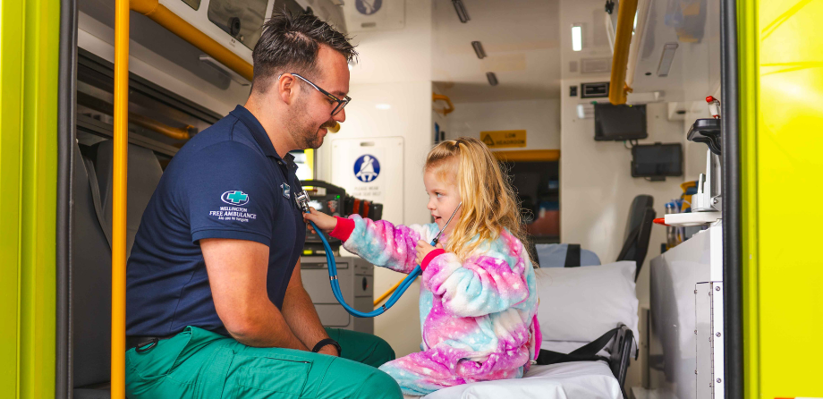 A paramedic in the back of an ambulance with a young child in a onesie, holding a stethoscope to their chest