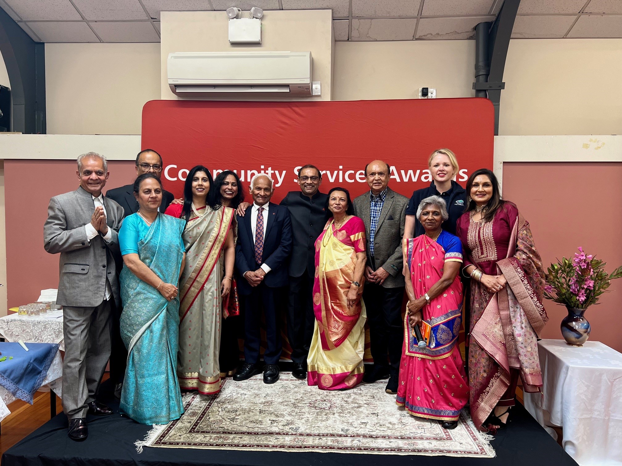 A group of Wellington Indian Association members stand in front of a red sign that says: Community Service Awards