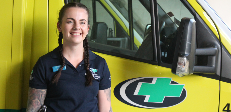 Kirsty has long dark brown plaits and is pictured wearing a dark blue Wellington Free Ambulance polo shirt in front of an ambulance